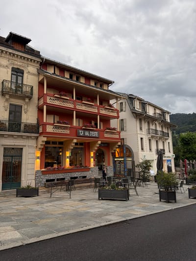 The image shows a street scene in Saint-Gervais-les-Bains, France, on an overcast day. In the foreground, a paved square with stone tiles is visible, with a dark asphalt road bordering it. Several dark planters with greenery and flowers are placed on the square, as well as outdoor seating areas with tables and chairs, some of which are covered with umbrellas.
Dominating the midground are two buildings. The building on the left is a light-colored, ornate structure with decorative stone carvings around a dark-windowed entrance and a balcony. Adjacent to it is a prominent red building with multiple balconies featuring decorative white railings and wooden shutters. The red building has a sign that reads "VAL D'ESTE" above its ground-floor windows, which appear to belong to a restaurant or cafe.
To the right of the red building is a taller, white building with a sloped dark roof. This building also features balconies with dark railings and numerous windows. Signs for "CREDIT AGRICOLE" and "RESTAURANT" are visible on this building and its ground floor.
In the background, a wooded mountainside is visible under the cloudy sky, suggesting a location in a mountainous region. A single person is walking on the pavement in front of the red building. The overall impression is of a European town square, likely a commercial area with dining establishments.