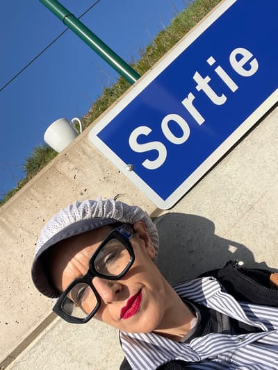 The image depicts a woman posing outdoors, illuminated by direct sunlight, against a concrete structure in Roodt-sur-Syre, Luxembourg. The perspective is a low-angle selfie, rotated counter-clockwise.
The woman occupies the lower-left and central portions of the frame, looking upward and slightly to her right, with a closed-mouth smile. She wears large, dark-framed eyeglasses and bright red lipstick. Her head is covered by a light-colored, possibly white or off-white, textured or ruffled cap/bonnet. She is dressed in a striped shirt, visible beneath a dark, possibly zippered, garment. Strong shadows are cast on her face and the concrete surface, indicating bright daylight.
Above and to the right of the woman is a blue rectangular sign with a white border, featuring the word "Sortie" (Exit) in large white capital letters. The sign is attached to a concrete surface with two visible silver fasteners. To the left of the sign, resting on a concrete ledge, is a plain white ceramic mug with a handle facing right. Behind the concrete structure, a grassy embankment slopes upward, and a green cylindrical pipe or pole is visible against a clear blue sky.