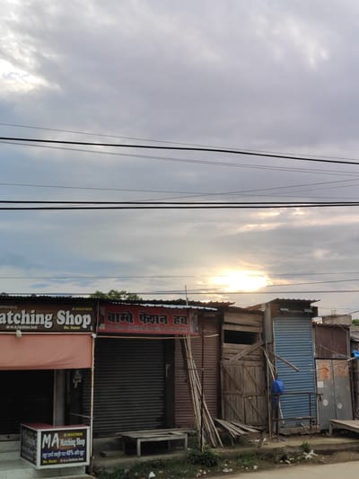 The image depicts a street-level view of a commercial area in Sitarganj, India, with a focus on two adjacent shops. The sky above is partly cloudy with the sun peeking through, suggesting it might be late afternoon or early evening. Overhead power lines are prominently visible, running horizontally across the frame.
The shop on the left is labeled "MA Matching Shop" and features a prominent sign with the shop's name in English and a smaller Hindi text indicating a discount. Below this sign, a display box showcases some items. The facade of this shop is a light pinkish color with a dark awning. The shop to its right has a sign in Hindi that reads "बाल्मी फैशन हब" and below it, "ALL GIRLS FASHION AND GENTS PENT SHIRT" in English. This shop has a dark, possibly metal, shuttered entrance.
The buildings appear to be constructed with a mix of materials, including corrugated metal and wood, giving them a somewhat makeshift or rustic appearance. Several wooden poles and planks are stacked outside the right shop. A blue plastic container is visible near the entrance of the right shop. The ground in front of the shops is a dirt path with some scattered debris, including plastic bottles and wrappers. The overall environment suggests a bustling, possibly developing, commercial zone. No people are visible in the frame.