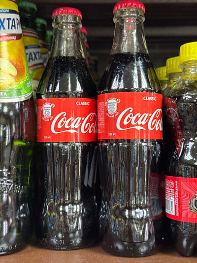 The image shows a close-up of several glass bottles stacked together, likely on a store shelf. The main focus is on two bottles of Coca-Cola Classic, easily identifiable by their red labels with the white Coca-Cola logo and the "CLASSIC" designation. These bottles have red caps and are filled with a dark liquid. Below the Coca-Cola bottles, a green and yellow bottle is visible with the Russian word "ΧΤΑΡΙ" written on it. The top edge of another Coca-Cola bottle is visible with a yellow bottle nearby, and its cap color is yellow. The text on the Coca-Cola bottles, though partially obscured, includes nutritional information and volume (330 ml). The Coca-Cola bottles also have a barcode and some Russian text that includes the words "ЭТА БУТЫЛКА ПЕРЕРАБАТЫВАЕМАЯ" which translated to "This bottle is recyclable". The lighting suggests an indoor environment, possibly a store or warehouse setting. The exact location within Chuprino, Russia, cannot be determined from the image alone.