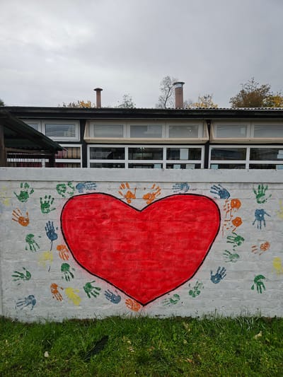 A close-up, exterior shot shows a white brick wall painted with a large red heart outlined in black. Around the heart are colorful handprints in shades of blue, green, orange, and yellow. The wall abuts a building with white-framed windows. To the right of the wall is a patch of green grass. The photograph is taken in Osorno, Chile.