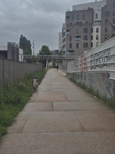 The image captures a street scene in Aubervilliers, France, characterized by modern architecture and urban elements. A sidewalk, composed of large square pavers, dominates the right side of the frame, bordered by a strip of grass. A trash can sits on the grass next to the sidewalk. A fence runs parallel to the sidewalk, separating it from a lower area with trees and a pedestrian overpass. On the left, tall apartment buildings with multiple windows rise into a gray, overcast sky. A corrugated metal structure overhead is covered in graffiti. In the distance, a figure is walking on the sidewalk.