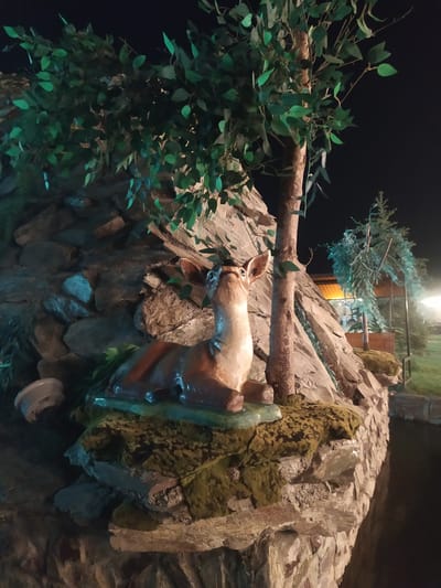 This is a night-time, medium-shot, low-angle photograph of a decorative scene featuring a life-sized statue of a deer resting on a moss-covered rock formation. The deer statue is light brown with dark brown accents and is looking upwards with its head tilted. A thin, artificial tree with green leaves rises behind the deer. The rock formation is constructed from various sizes of stones and appears to be part of a larger, possibly artificial, landscape. In the background, a small, artificial evergreen tree and a building with illuminated windows are visible. The overall setting suggests an outdoor decorative display, possibly in a park or a private garden, in the city of Sosnovka, Kyrgyzstan. The lighting is artificial and casts shadows, creating a dramatic atmosphere.