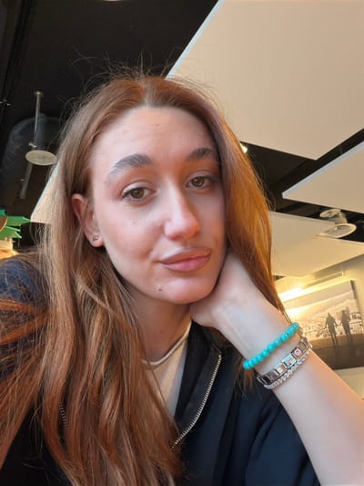 A close-up, rotated image shows a woman with fair skin and auburn hair resting her left hand at her neck. She is wearing three bracelets on her left wrist, including a blue beaded bracelet and two metal bracelets. She has light brown eyes and is wearing a small earring in her left ear. She is wearing a black jacket over a white top. The background is a mix of neutral tones, including a white ceiling with black geometric patterns, as well as a picture frame. The location of the image, Lucerne, Switzerland, cannot be determined from the visual content.