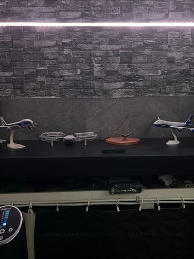 This is a forensic, matter-of-fact description of a scene, located in Schoonderbuken, Belgium.

The image depicts a shelf against a dark, textured wall, possibly wallpaper designed to resemble brick. A strip of LED lights illuminates the top portion of the wall. On the shelf, several objects are arranged. To the left, a model airplane with white wings and a blue fuselage is displayed on a white stand. Next to it, a small white drone with protective cages around its propellers is positioned. Further to the right, a small, ornate brass lamp, resembling a genie lamp, sits on a red, heart-shaped wooden base. An incense stick is inserted into the lamp, with smoke rising from its tip. To the far right, another model airplane, similar in design to the first but with "SILKWAY" written on its fuselage, is also on a white stand. Below the shelf, the image shows the top of a white unit with various wires and clips attached to it, and a black patterned surface below that. On the far left, a circular device with a digital display and buttons is visible, labeled "QuietSet". The overall environment suggests an indoor setting, possibly a display or storage area.