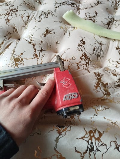 The image captures a close-up view of a hand holding a red and silver stapler gun, positioned against a light-colored, quilted surface.
The central focus is a person's hand, partially visible on the left side of the frame, holding a red-bodied stapler gun with a silver magazine housing. The hand's fingers are wrapped around the handle of the tool, with the index finger resting near the trigger. The stapler gun's metallic nozzle is pressed against the soft, tufted surface.
The background consists of an upholstered or quilted fabric, which is a light, creamy beige or off-white color. This fabric is adorned with prominent, irregular, and wavy patterns in a metallic gold or bronze hue, resembling a marble or veined texture. The surface also exhibits a quilted or tufted design, characterized by depressions and raised sections, suggesting it might be a headboard, a large cushion, or a mattress.
In the upper right corner of the image, a light green, slightly curved piece of porous material, likely foam or sponge, is visible. It appears to be a cut strip or section of foam.
The overall scene suggests an activity related to upholstery, crafting, or repair, where the stapler gun is being used to attach fabric or materials to the quilted surface.
Visible text on the red body of the stapler gun clearly shows "CYCO" in silver lettering, with another stylized symbol (possibly an intertwined "S" or a logo) above it. The surface of the stapler gun appears somewhat worn, with minor scratches and scuffs.