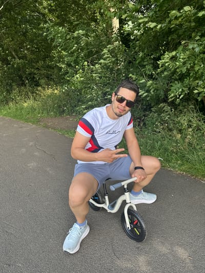 A man is posing for a photo while straddling a small child's bicycle on an asphalt path. He is wearing sunglasses, a white t-shirt with red and blue stripes on the sleeves, grey shorts, and light blue athletic shoes. The man is making a peace sign with his left hand. The background consists of lush green trees and foliage. The setting appears to be a park or natural area. This scene is not identifiable as being in the city of Wakefield, United Kingdom.