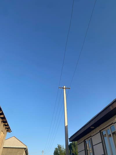 In the city of Боғистон, Tajikistan, a concrete utility pole stands tall against a clear blue sky. Three dark electrical wires stretch diagonally across the upper portion of the frame, originating from the top of the pole and extending upwards and to the right. On the left side of the image, the roofline of a building is visible, with exposed wooden rafters and a gray corrugated roof. To the right of the utility pole, another building is depicted, featuring a dark brown, corrugated roof overhang above a light-colored wall with dark horizontal decorative bands. Multiple windows with reflective glass are present on this building's facade. The lower right corner of the image shows the tops of green trees.