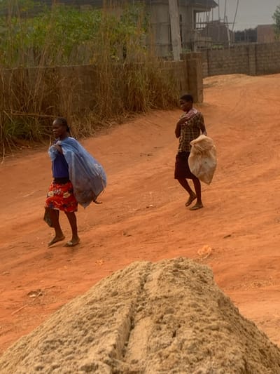 Two individuals traverse a dirt path, carrying what appear to be sacks on their backs. The path is flanked by vegetation on one side and a large, textured object on the other. One person dons a plaid shirt and dark shorts, while the other wears a blue shirt and a red floral skirt. The backdrop features buildings in varying stages of construction, under a light sky.