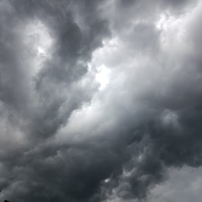 A sky filled with ominous, dark storm clouds dominates the frame. Various shades of gray and black swirl together, indicating a potentially severe weather event. A small patch of bright, white light appears amidst the dense cloud cover, hinting at the sun attempting to break through. The clouds are thick and heavy, creating a sense of impending drama and atmospheric tension.