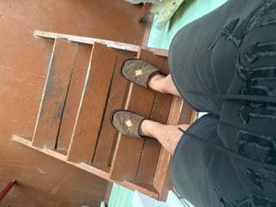 A person is standing on wooden stairs wearing brown slippers with a diamond-shaped pattern on them and dark shorts. The stairs are painted brown and appear worn. The environment suggests an indoor or outdoor setting with a light green floor visible in the background. The camera angle is from above, looking down at the person's legs and feet. There is no indication of the specific location in Krasnoe, Russia.