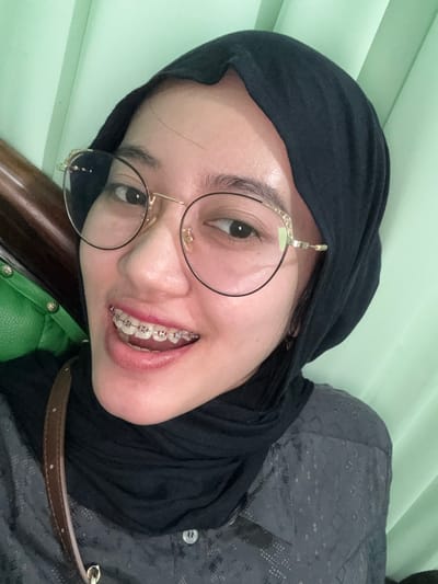The scene is an indoor setting, identified as being in Patrang, Indonesia. A single individual, presumed female, is positioned facing the camera, smiling directly into the lens. The individual wears a black hijab covering the head and neck, and large, round spectacles with thin gold-colored frames. Dental braces are visible on the upper teeth, marked by silver brackets and a pink band. The individual is wearing a dark grey long-sleeved shirt with a subtle patterned texture. A brown strap, indicative of a shoulder bag, is visible across the individual's right shoulder. The background predominantly consists of a plain light green surface, possibly a wall or curtain. To the left of the individual, a dark brown object and a portion of a green cushioned surface, possibly part of a chair or sofa, are discernible.