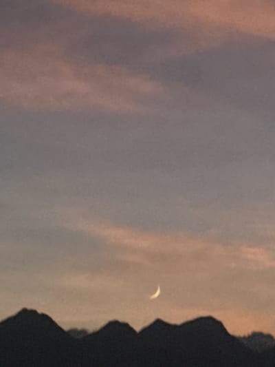 The image depicts a natural landscape at twilight, observed from Las Veredas, Mexico. The upper two-thirds of the frame show a sky with a gradient transition from a muted purplish-gray at the top to a softer orange-pink hue towards the horizon. Diffuse, indistinct cloud formations are present across the sky. A slender, illuminated crescent moon, appearing white or pale yellow, is visible in the lower-middle section of the sky, slightly right of the vertical center. The lower third of the image is dominated by a dark, silhouetted mountain range with irregular peaks and valleys, extending horizontally across the entire frame. No human subjects, animals, or man-made objects are visible, and no actions or interactions are depicted. The environment is entirely natural.