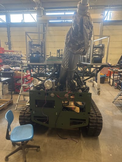 The image presents an interior view of an industrial workshop located in Logatec, Slovenia. Centrally positioned is a large, olive-green tracked robotic platform, appearing to be under assembly or maintenance. Its chassis is partially exposed, revealing internal components such as electric motors, complex wiring harnesses, and exposed circuit connections with red and black alligator clips attached to the front panel. A substantial silver-colored cylindrical mast, partially enveloped in crumpled grey protective plastic sheeting secured by a metal chain, extends upwards from the platform, angled towards the upper left of the frame. The platform rests on what appears to be a wooden pallet.

To the immediate left foreground, a light blue office chair with a perforated seat, backrest, and five-wheeled base is situated. The workshop floor is grey concrete. The background reveals an active industrial environment: to the left, cluttered metal shelving, red multi-drawer tool carts, and various components and scrap materials are arranged. A two-step metal ladder is visible adjacent to a tool cart. To the right, sections of what appear to be machinery or an assembly line with a green conveyor belt are visible, along with a mobile metal frame structure on wheels. Overhead, a yellow gantry crane system, labeled with "2000 KG" and "J.MIHO," is installed. Large, high-set windows line the distant wall, providing natural illumination. No individuals are present in the scene.