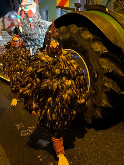 The image displays a nighttime street scene in Wasserbillig, Luxembourg. Two individuals are present, dressed in elaborate chicken-themed costumes. The person in the foreground wears a dark brown feathered suit with a rooster headpiece featuring a red comb, yellow beak, and small white lights embedded on the head and torso. Orange fabric wraps the lower legs, terminating in yellow webbed feet. To the left and slightly behind, a second individual wears a similar dark feathered suit with a red and yellow chicken headpiece that exposes their face; they also have yellow webbed feet. Both are positioned on a dark, wet-looking asphalt surface, which is littered with scattered confetti or small paper debris. A large green tractor occupies the right side of the frame, its prominent black, deeply-treaded rear tire dominating the view, with a red and yellow reflective hazard marker visible on its upper frame. In the background, behind the costumed figures, a large, illuminated chicken-shaped parade float with multi-colored appendages is visible, situated in front of a light green building.
