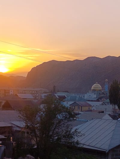 The image depicts a sunset over the city of Naryn, Kyrgyzstan, with a prominent mountain range in the background. The sky transitions from a bright yellow and orange near the horizon, where the sun is setting, to a soft pink and peach hue higher up. Several power lines are visible in the sky, silhouetted against the vibrant sunset.

In the midground, a cluster of buildings forms the cityscape. Among them, a mosque stands out with its golden dome and a tall, slender minaret that is blue and white. The mosque is adorned with intricate patterns. Surrounding the mosque are residential buildings with varying roof types, including some with corrugated metal roofing and others with lighter-colored materials. Trees are scattered throughout the city, with some appearing in the foreground as dark silhouettes against the illuminated sky. The overall impression is of a serene evening scene in a mountainous urban environment.