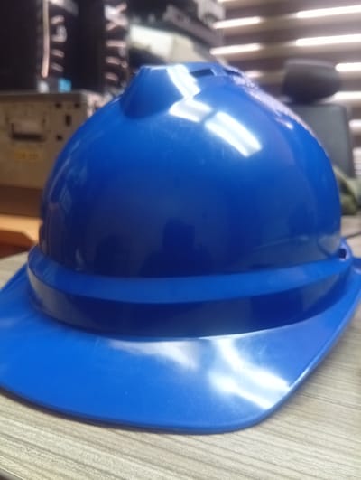 A bright blue hard hat with a wide brim sits on a wooden table. The hat has a rounded crown with ventilation holes and a raised ridge around the base of the crown. The background is blurred, showing what appears to be an indoor environment with horizontal blinds and some objects on shelves. The lighting in the scene creates bright reflections on the shiny surface of the hard hat. There are no people visible in the image. The location is identified as Shiroro, Nigeria.