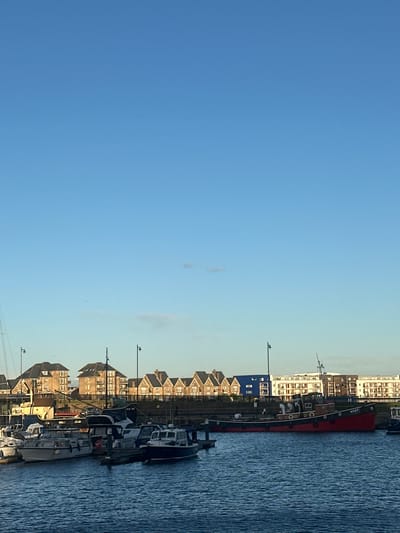 The image depicts a marina scene within the city of Gillingham, United Kingdom, under a clear, pale blue sky with minimal, wispy cloud cover. The foreground is dominated by calm, dark blue water with slight ripples.
Moored in the water are several boats. On the left and central-left, a cluster of white and light-colored leisure craft, including motor yachts and cabin cruisers, are docked at dark-colored pontoons. These vessels feature enclosed cabins, railings, and various antennae. To the right, a prominent, older-style vessel, identified by the name "KENT" on its stern, is moored. This boat has a dark red hull, a black upper structure, and circular portholes along its side. Further right, additional smaller pleasure boats with dark blue hulls are visible. Dark metal lampposts are positioned along the pontoons and waterfront.
The background features a built environment. On the left-middle, a series of multi-story residential buildings with traditional pitched, dark tiled roofs and brown or beige facades are visible. To the right of these, a modern multi-story building with a prominent bright blue section is present. Adjacent to the blue building, a longer, multi-story structure with a lighter, possibly white or cream-colored facade and numerous windows completes the background architecture. The buildings are situated behind a barrier or on slightly elevated ground overlooking the marina. The scene is illuminated by natural light, casting subtle shadows consistent with a low sun angle. No individuals are visible, and no specific actions or interactions are depicted.