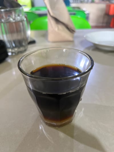 In Manyang Cut, Indonesia, a close-up, eye-level shot captures a clear glass tumbler filled with dark liquid, possibly coffee or tea, resting on a light-colored, smooth surface. The beverage has a frothy layer at the top and a sediment at the bottom. The glass exhibits a faceted design, and light reflects off its surface. In the background, out of focus, are various objects including a stack of papers or a bag, a clear jug containing water, and glimpses of green and red objects, possibly furniture or containers. The overall setting appears to be an indoor environment, possibly a cafe or a home dining area.