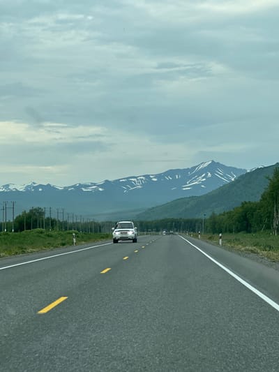 An eye-level, point-of-view shot depicts a car driving on a two-lane asphalt road, which stretches into the distance. The road is marked with solid white lines on the right and dashed yellow lines on the left, indicating traffic flow. The sky above is overcast with light gray and white clouds. In the middle ground, on either side of the road, there is a grassy shoulder lined with utility poles. Further back, a dense line of green trees forms a backdrop, beyond which lie majestic mountains with snow-capped peaks. A white SUV is visible on the road, driving towards the viewer. There are no people visible in the image. The scene appears to be set in a rural or mountainous area, possibly in the vicinity of Кеткино, Russia, given the presence of utility poles and the natural landscape.