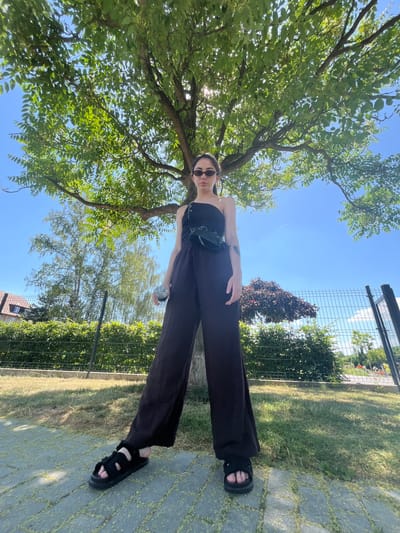 A young woman stands outdoors on a sunny day, posing under a tree with bright green leaves. She is wearing a black tank top, black wide-leg pants, and black fuzzy slide sandals. A black fanny pack is worn across her chest. She is also holding a clear glass water bottle in her right hand. The woman is wearing dark sunglasses.

The background shows a lush green lawn and a fence. The sky is clear and blue with a few wispy clouds. The sun is shining brightly, casting shadows. The scene appears to be a park or a backyard, given the presence of the tree, lawn, and fence. The overall mood is relaxed and casual, suggesting a warm, pleasant day. The time of day appears to be midday, given the bright sunlight and high angle of the sun. There is no visible text in the image.