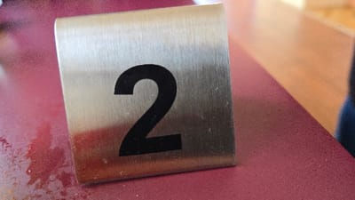 A metal table number displaying a large, black "2" sits on a maroon tabletop. The metal has a brushed texture. Parts of the table surface appear slightly wet, with darker patches visible near the table number. The blurred background shows hints of a light-colored wall and a person's arm.