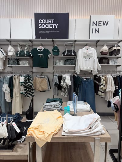 This media file captures a retail display of clothing and accessories, likely within a store in Dagupan, Philippines. The scene is an interior shot of a clothing store, organized with shelves and racks showcasing various apparel. Dominating the left side of the image is a large white sign with the bold, dark text "NEW IN". Below this, another sign reads "COURT SOCIETY" with a tagline suggesting a collection of "pleated silhouettes with subtle sport-inspired accents."
The merchandise displayed includes a variety of garments such as t-shirts, hoodies, jeans, shorts, and hats. Prominent among the items are a white PENSHOPPE hoodie, a dark green t-shirt with "THE CREW" text, and several stacks of striped t-shirts and solid-colored t-shirts in white, cream, and a pale yellow. There are also several pairs of jeans and folded shorts. Accessories like black duffel bags and caps are visible.
The overall impression is one of a well-stocked clothing store with a focus on casual and sporty wear. The lighting suggests an indoor retail environment, possibly during daytime hours given the typical operation of such establishments. There are no people visible in the image, focusing the viewer's attention entirely on the products and store layout. The arrangement of items is neat and organized, indicating a deliberate presentation for customers.