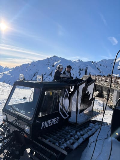 An outdoor DJ event is depicted on a snow-covered mountain in the vicinity of Schwendau, Austria. Two individuals are positioned atop a modified black tracked vehicle, resembling a snowcat, which functions as a mobile DJ booth. The person on the left, wearing a light-colored beanie, operates a DJ mixer and turntables. To their right, a second individual in a dark baseball cap and dark attire observes. The vehicle is branded with "PHIERES" on its side and "phieres.com" on the windshield, which also displays "Schneekarhütte.at." Two spotlights are mounted on the cab roof. Large black and white speaker enclosures or decorative panels, featuring a stylized mountain logo, extend from the rear of the DJ setup. The background features extensive snow-covered mountains under a clear, bright blue sky, with a visible sun in the upper left. A distant peak exhibits a ski lift station. To the right of the scene, a multi-level building constructed of stone and wood, featuring a wooden bridge or walkway, is visible, with several individuals present, indicating a ski resort or mountain lodge environment.