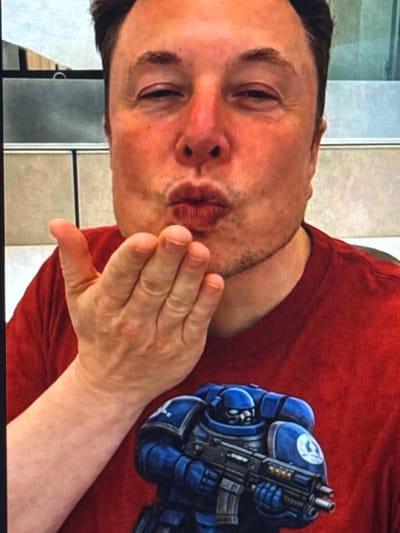 The image shows a close-up of a man wearing a red t-shirt with a blue Warhammer 40,000 Space Marine on it. The man is blowing a kiss, with his hand held up to his lips. He has dark hair and light skin. The background is blurry, but appears to be an indoor setting with some vertical blinds or partitions. There is no information in the image or provided context that indicates this scene is located in Shcherbanka, Ukraine.
