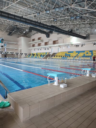 A large, modern indoor swimming pool facility is depicted, likely located in Tiraspol City Council, Moldova. The pool itself is deep blue, divided into lanes by white and red ropes. Several lane markers, including triangular flags with the "Arena" logo and numbered starting blocks (8 and 7 are visible), line the pool's edge. Spectator seating is arranged in tiered rows behind the pool, with chairs in alternating yellow and blue colors. The ceiling features an extensive metal truss system and industrial lighting fixtures, indicating a substantial construction. The surrounding area is tiled, and green footwear is placed near the pool's edge. A single individual, wearing a red shirt, stands on the far side of the pool.