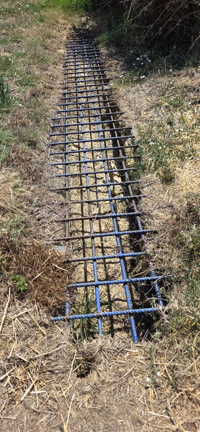The image shows a long, rectangular grid of metal rebar, likely for construction or reinforcement, laid out on the ground. The rebar is dark gray and has a twisted texture, and some sections appear to be painted blue. The grid is oriented horizontally across the frame and extends into the distance.

The setting is outdoors, in a rural or natural environment. The ground is covered with dry, straw-like vegetation and some patches of green grass. There are also some scattered small white wildflowers visible in the dry grass. To the left of the rebar grid, there is an area of darker, more dense vegetation, possibly bushes or thorny plants, with some of the same small white flowers.

The time of day appears to be daytime, suggested by the bright, directional shadows cast by the rebar onto the ground. The lighting indicates a sunny day. There are no people visible in the image, and no specific activity or event is clearly taking place beyond the rebar being laid out. The overall impression is one of preparation or construction in a natural setting.

The location context provided is Angüés, Spain, which is consistent with the dry, Mediterranean-like vegetation. No text is visible in the image.