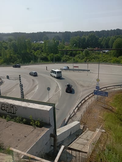 The image depicts a view of a highway intersection in Koltsovo, Russia, on a sunny day. A white minibus and a dark-colored hatchback are visible on the road. A person stands near the intersection. The environment is characterized by a paved road with lane markings, guardrails, and vegetation on the surrounding hillsides. Signage at the intersection indicates directions to "Koltsovo" and "Orlovka." In the foreground, a concrete structure with graffiti is present, suggesting an elevated vantage point. The sky is clear.