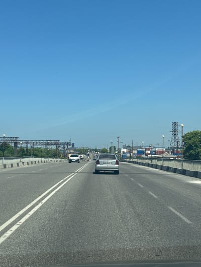 A bright, clear blue sky is visible above a multi-lane asphalt road. The road appears to be on an elevated structure, possibly a bridge or overpass, with a concrete barrier lining the right side and a metal railing along the left side. Several cars are on the road, traveling in the same direction. The car in the foreground, on the right, is silver with a visible license plate. To the left, a white car is further ahead. In the distance, industrial structures, including large metal frames and shipping containers, are visible, suggesting proximity to a port or industrial area. Green trees line the left side of the road in the background. Power lines and utility poles are also present. The overall scene depicts a sunny day in an urban or industrial setting, likely the city of Poti, Georgia, given its known port and industrial activities.