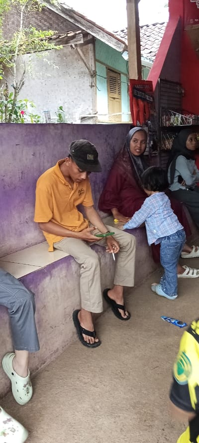 The image shows a scene in Cimaung, Indonesia, at coordinates -7.074 latitude, 107.547 longitude. A man is sitting on a bench, wearing a yellow shirt and beige pants. He is holding a cigarette and looking down. He is wearing black flip-flops.  A woman wearing a red headscarf is sitting to the right of the man. A young child in a blue and white plaid shirt is standing beside the woman, looking towards the camera. Another woman is sitting behind the child, wearing a blue shirt and a black headscarf. She is looking away from the camera. They are all sitting or standing in front of a wall painted a light purple color.  The scene is in front of a building with a pale blue wall and an open doorway.  The ground is covered with tan colored concrete. There is a partially visible pack of cigarettes on the ground.  The image is captured from a medium distance.  The scene is outdoors.  The weather is sunny. The sun is not directly in the image.  The scene is calm.  The subject of the image is the man sitting on the bench.