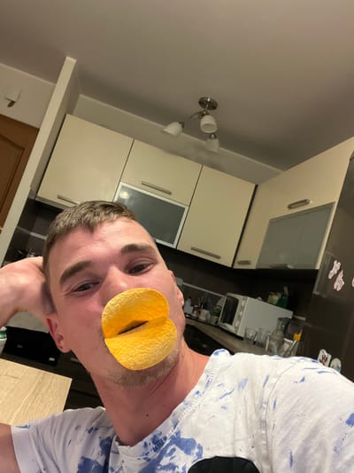 A selfie is taken in what appears to be a kitchen in a residential apartment in the city of Rumia, Poland. The subject, a Caucasian male with short brown hair, is positioned in the foreground, with his head tilted back and his left arm bent, his hand resting behind his head. He is wearing a white t-shirt with blue paint splatters. Two potato chips are covering his mouth, giving the appearance of lips. The background features kitchen cabinets with cream-colored doors and stainless steel handles, and a dark countertop with various kitchen appliances and items, including a white microwave. The lighting suggests an indoor environment, likely during the evening.