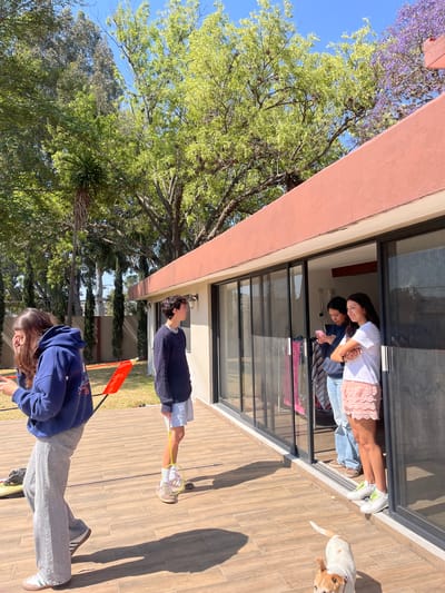 Two women are inside a building looking out of a window. The location is El Oasis Valsequillo, Mexico. The scene depicts the exterior of a building with two women and a dog inside, looking out of a large window. Outside, two other people are visible: one standing, holding what appears to be a badminton racket, and another bending over. The overall environment includes trees and clear sky.