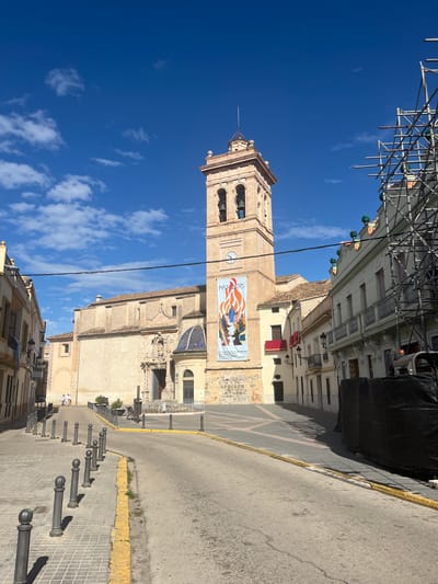 The image shows a street view of the historic center of Torrent, Spain, featuring the Church of the Assumption of Our Lady. The church, made of beige stone with a tall bell tower, is adorned with a large banner depicting a scene with multiple people. The clear blue sky has scattered clouds. Scaffolding is visible in the upper section of the frame. The street has a yellow curb and is lined with bollards. Buildings are seen on either side of the street, with awnings visible on one side. Power lines are crossing the sky and the top of the church.