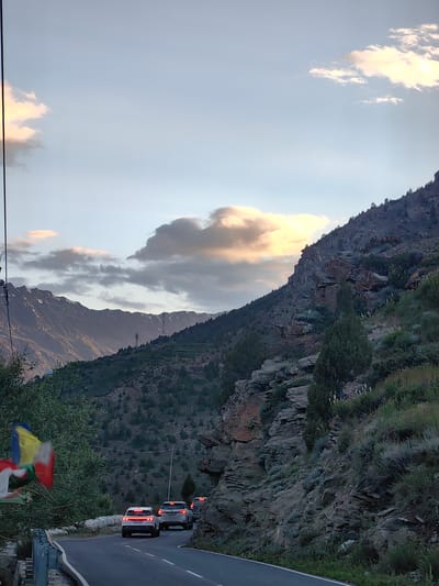 This image captures a scenic mountain road in Kead, India, at dusk. The setting is a winding asphalt road with white lane markings, bordered by a steep, rocky, and vegetated mountainside on the right. On the left, there is lush green foliage and a prayer flag, adding a touch of cultural context. In the distance, snow-capped mountains rise majestically against a sky filled with dramatic, colorful clouds illuminated by the setting sun.
Several vehicles are present on the road, suggesting a journey or commute. A white SUV is in the foreground, followed by another white SUV, and a dark-colored vehicle further back, all traveling in the same direction. The headlights and taillights of these vehicles are visible, indicating the fading light of evening.
The time of day is clearly indicated by the warm, golden light hitting the clouds and the softening of the landscape's colors. The weather appears to be fair, with scattered clouds adding visual interest to the sky. The overall mood conveyed is one of peaceful travel through a breathtaking natural landscape. The presence of the prayer flag hints at a spiritual or cultural aspect of the location. No visible text is present other than the license plate on the white SUV, which appears to read "PB19 J 8694".