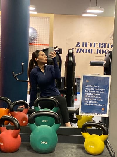 This is a vertical, eye-level shot taken inside a gym. The foreground is dominated by a row of kettlebells in various colors, including red, green, and yellow, with weights marked in kilograms: 8 KG, 20 KG, 45 KG, and 2 KG. In the mid-ground, a woman with dark hair and white earbuds is taking a selfie with her phone. She is wearing a dark blue athletic top and black leggings. Behind her, a weight machine and a sign with Swedish text are visible. The text on the sign, which appears to be gym instructions, reads: "Du får gärna använda de tyngre hantlarna här inne, men hantlarna tillhör allmänna friviktsytan. Glöm inte att ställa tillbaka dem efter er!" (English translation: "You are welcome to use the heavier dumbbells in here, but the dumbbells belong to the common free weight area. Don't forget to put them back afterwards!"). The background features a large graphic on the wall with the words "STRETCH" and "DOT DOWN" in large, bold lettering. The gym setting is implied by the equipment and the signage. The lighting appears to be artificial, typical of an indoor gym. No specific time of day or weather conditions can be inferred. The woman's expression is neutral, focused on taking the selfie.