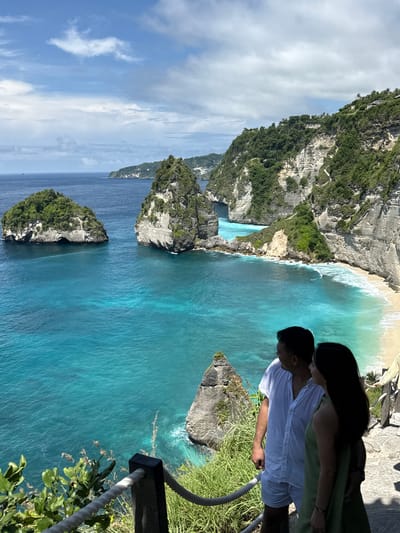 The image shows a couple standing on a cliff overlooking a beach. The woman is wearing a green dress, and the man is wearing a white shirt and shorts. They are both looking out at the ocean. The cliff is covered in green vegetation, and the beach is sandy with turquoise water. There are several small islands in the distance. The location is Nusa Penida, Pejukutan, Indonesia. There's a fence nearby on the cliffside.