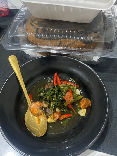 The image displays an overhead view of a meal setting indoors. In the foreground, a round, matte black bowl contains a dark-sauced dish. Visible ingredients in the bowl include vibrant green leafy vegetables, several whole red chili peppers, sliced white garlic cloves, and chopped red tomatoes. A gold-colored spoon rests partially submerged in the dish, holding a single cooked, orange-pink shrimp. The bowl is positioned on a dark gray or black fabric surface, which appears to be a placemat, with a white surface visible to the lower right.
In the background, two stacked food containers are present. The top container is a white, opaque foam or plastic takeout box lid, partially obscuring the item below. Beneath it, a clear plastic clamshell container holds a round, baked good, possibly a cake or bread, featuring a distinct dark brown swirl pattern on a lighter brown base. This baked good rests on a white, lacy paper doily. A translucent red cup or glass is partially visible on the far left in the background. The specific location of the scene in the city of Cibinong, Indonesia, cannot be visually confirmed from the provided image.