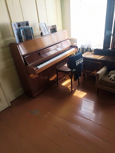 The image depicts an interior scene with a piano. A dark blue t-shirt with white Cyrillic lettering is draped over a small wooden stool placed in front of the piano. The piano itself is an upright model with a wooden finish. On top of the piano, several framed photographs and what appear to be printed photo albums or books are arranged. In the background, a sheer white curtain covers a window, and to the right, a dark wooden dresser or cabinet is visible. A light-colored armchair with a stuffed animal on it is also present in the right side of the frame. The floor is made of wooden planks with a reddish-brown finish, showing signs of wear and tear. Sunlight streams in from the window, casting shadows on the floor and the piano. This scene is located in Aygezard, Armenia.