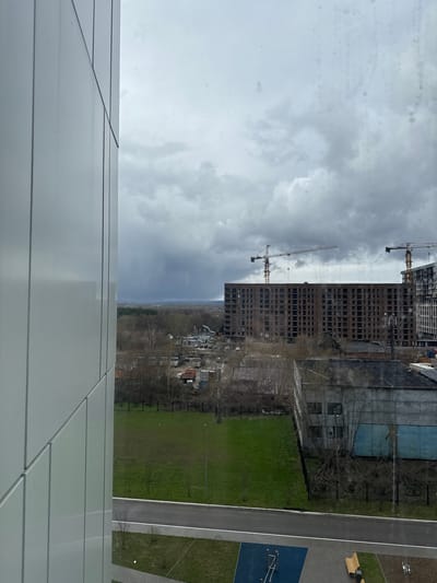 The image presents an outdoor scene viewed from an elevated perspective, encompassing urban development and natural landscapes under an overcast sky. In the immediate foreground, the light gray, paneled exterior of a modern building, from which the photograph was taken, occupies the left and bottom portions of the frame.
The midground is dominated by two large, multi-story buildings under construction, featuring brick and concrete skeletal structures with numerous window openings. Two tall tower cranes are prominent beside these buildings, indicating ongoing development. To the right, a paved road or pathway runs diagonally, separating sections of vibrant green grass. Adjacent to the path is a children's play area with a blue surface and white structures, and a wooden bench. Further back, another green field with bare trees and a metal fence borders an older, possibly industrial-style building with a partially blue facade and visible windows.
In the background, a dense line of deciduous trees, mostly bare, forms a forest, behind which a cluster of smaller buildings or houses is visible at the horizon. The sky is uniformly cloudy and overcast, creating a soft, diffused light across the scene. The overall impression is one of a developing area, blending new construction with existing infrastructure and natural elements. No distinct text is visible in the image. The scene depicts a daytime view in what appears to be a suburban or semi-rural setting, consistent with the location context of Borovoye Matyushino, Russia.