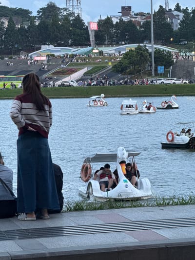 The image captures a scene at Xuan Huong Lake in Da Lat, Vietnam. In the foreground, a woman with dark hair, wearing a striped sweater and a long denim skirt, stands with her back to the camera, her hand resting on her hip. To her left, another person, partially visible, is seated. The woman is wearing Crocs.
The lake is populated with several swan-shaped paddle boats. One boat in the mid-ground carries at least three people. Another, further back, also has passengers. To the right, a larger boat with a canopy is seen, with people visible inside. In the foreground, a swan boat is partially visible, with two men wearing life vests seated in it.
In the background, a grassy bank slopes up towards a series of tiered steps. Numerous people are scattered across these steps and the grassy areas. A large green bus is visible on the left side of the frame. Further up the hillside, buildings and trees are visible, and a large tower with a red "GO!" sign stands out against the sky. Cars and motorcycles are parked and moving along a road in the mid-background. The sky appears overcast.