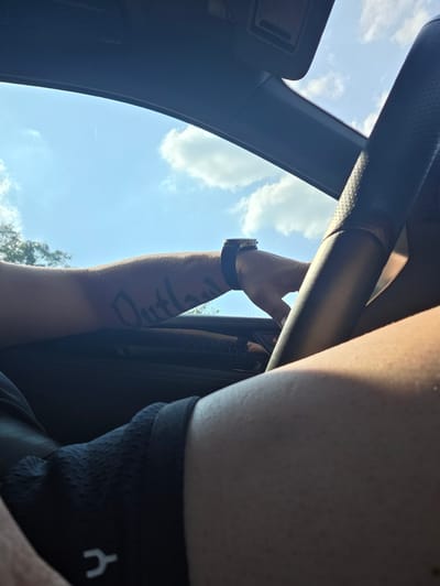The image shows a close-up view from inside a vehicle looking outwards through an open sunroof. The sky is bright blue with scattered white clouds, suggesting it is daytime with clear weather. Sunlight is filtering into the car, casting shadows and illuminating parts of the interior and the occupant.

The visible subjects include parts of a person. We see a portion of their arm with a tattoo that appears to say "Outlaw" written vertically in dark ink. The person is wearing a black t-shirt or athletic top with a white symbol on the left shoulder area, and a black smartwatch on their wrist. Their hand is visible, resting on a dark-colored bar, likely part of the car's interior structure. Another part of the person's leg, possibly a thigh, is also visible in the foreground, suggesting they are sitting in the driver or passenger seat.

The setting is clearly the interior of a car, with dark panels and textured surfaces indicative of automotive design. The open sunroof frames a view of the sky and distant trees, hinting at an outdoor environment. The angle of the shot suggests the camera is positioned low, looking up and out.

The activity appears to be a casual moment, perhaps a drive on a sunny day, with the occupant enjoying the open air. The overall mood is relaxed and summery. There are no overt emotional expressions visible, but the bright lighting and open sky suggest a positive, possibly adventurous or leisurely experience. The location context provided is Rüsselsheim am Main, Germany, which is a city known for its automotive industry, aligning with the car setting.