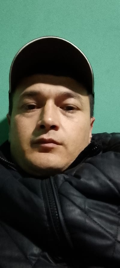 City of Tajikarab, Uzbekistan. A male individual, appearing to be in his 30s to 40s, is depicted in a close-up portrait, facing the camera directly with a neutral expression. He wears a dark baseball cap with a light or white brim and a dark, likely black, zippered puffer jacket. The lighting, possibly artificial, casts shadows under the cap brim, nose, and chin. The background is a plain, untextured mint green wall. No other individuals or objects are present.