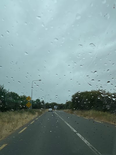 The image captures a view through a rain-streaked windshield on a cloudy day, looking down a wet asphalt road in The Row, Ireland. Trees line the sides of the road, and a yellow sign indicating a roundabout is visible on the left. A white car is driving in the distance. The overall atmosphere is overcast and damp, characteristic of a rainy day.