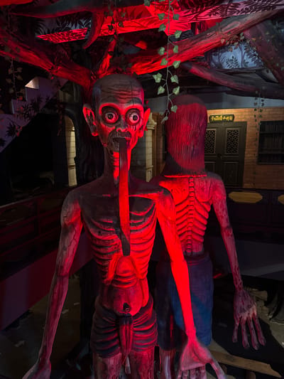 The image shows a distorted, red-lit scene, likely a display or installation, located in Bang Pu Subdistrict Municipality, Thailand. Two skeletal figures are prominently featured. One figure has an elongated face with exaggerated eyes and a stick protruding from its mouth. Its skeletal rib cage is visible. The other figure is positioned behind and above, also displaying a prominent rib cage. A sign written in Thai is visible in the background above the upper skeleton. The surrounding environment includes stylized trees and drapery, with red lighting bathing the entire scene. The scene appears to be theatrical or part of a themed environment.