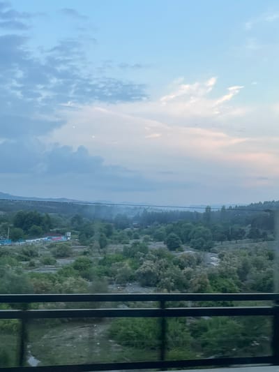 The image depicts a landscape scene captured from a vantage point behind a dark, vertical railing. The scene is oriented vertically, suggesting the photograph may have been taken on its side.

The left side of the image is dominated by a sky with scattered clouds. The clouds are a mix of white and light gray, with some areas appearing pinkish, indicative of a sunset or sunrise. The sky itself transitions from a light blue at the top to a softer, hazy hue closer to the horizon.

The right side of the image reveals a natural landscape with a body of water in the distance, possibly a lake or river, bordered by hills covered in dense greenery. The greenery consists of numerous trees and shrubs, creating a textured and somewhat rugged terrain. In the middle ground, there are some signs of human activity, including what appears to be a collection of buildings or structures with some red and white elements, possibly a small settlement or industrial area. A thin, dark horizontal line, likely a power line or cable, stretches across the landscape in the middle distance. The ground in the foreground appears to be a mix of dirt and vegetation.

The overall impression is that of a natural scene at dusk or dawn, with the soft light and hazy atmosphere contributing to a serene or perhaps melancholic mood. The railing in the foreground creates a sense of looking out from a structure, possibly a balcony or window. There is no discernible text in the image.