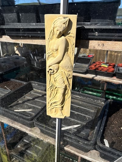 The image shows a rectangular, beige-colored bas-relief depicting a reclining female figure. The sculpture is placed on a greenhouse window, which reflects the surroundings, including potted plants and seed trays. The location appears to be in a garden. Shrewsbury, United Kingdom, is mentioned but is not visually confirmed in the image, and therefore cannot be incorporated into the verifiable description of the scene.